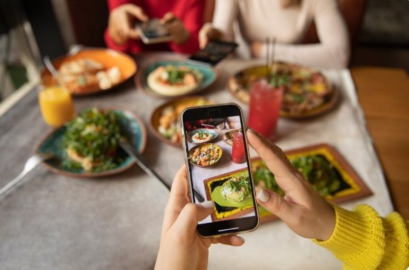 Person photographing colorful restaurant dishes on a smartphone for an Instagram food ad post.
