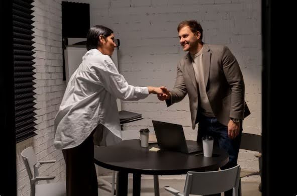 A woman in a white shirt and a man in a grey blazer shaking hands across a table with a laptop and coffee cups.