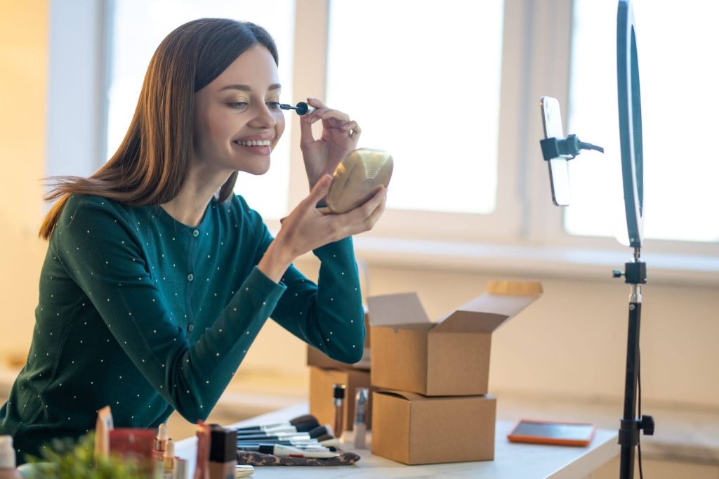A young woman in a green sweater applying mascara while filming herself with a ring light and smartphone setup.