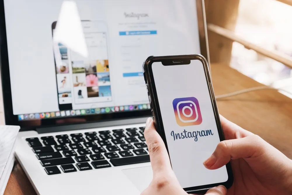 Hand holding a smartphone displaying the Instagram logo, with the Instagram login page open on a laptop in the background.
