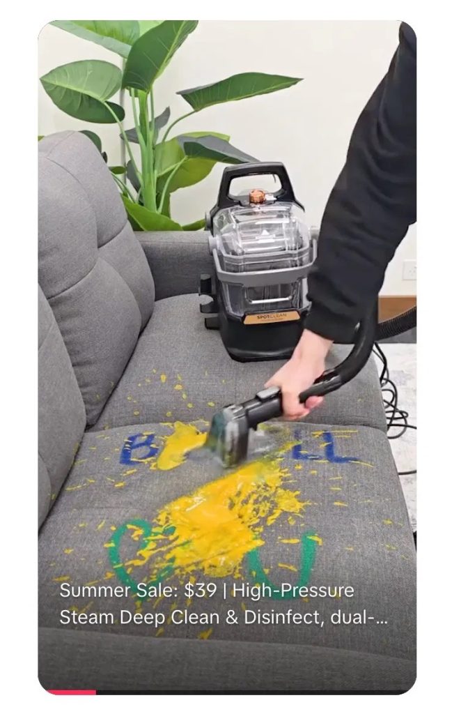 Person using a SpotClean portable carpet cleaner to lift yellow and blue paint stains from a grey fabric couch. Text overlay reads: "Summer Sale: $39 | High-Pressure Steam Deep Clean & Disinfect, dual-..."