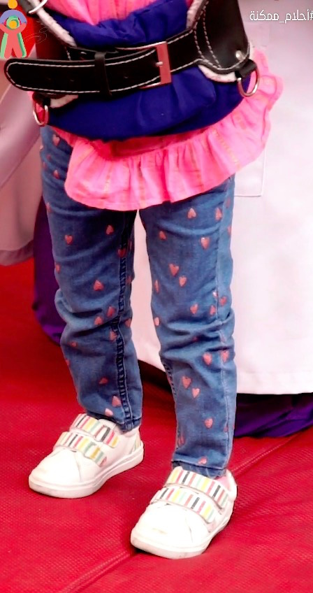 Child in heart-patterned jeans and white sneakers stands on red mat, wearing pink ruffled top.
