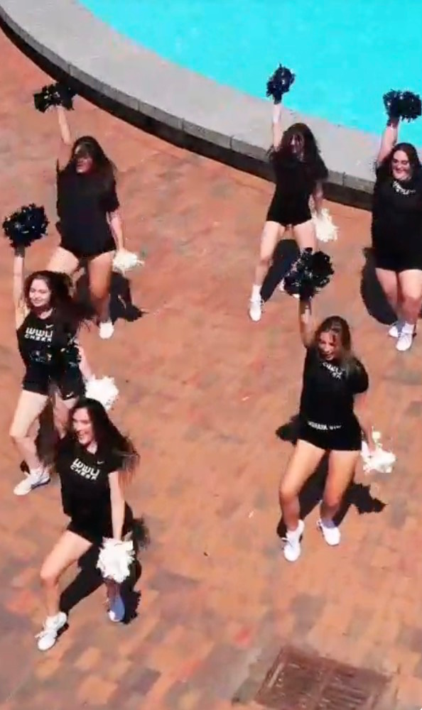 WWU cheer team in black uniforms performs with pom-poms on a brick plaza beside a pool, filmed from above.