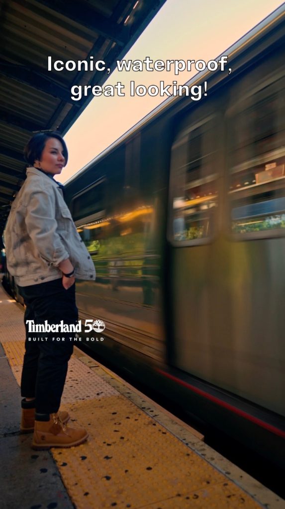 Woman in Timberland boots and cream jacket stands on a platform as a train speeds past.