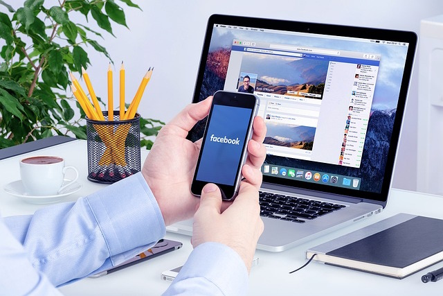 Person holding a smartphone displaying the Facebook app with a laptop showing Facebook in the background.