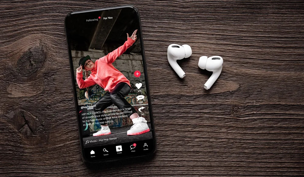 Smartphone displaying a TikTok hip hop dance video alongside wireless earbuds on a wooden surface.