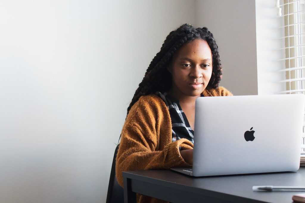 Woman in an orange cardigan working on a MacBook laptop at a desk near a window.